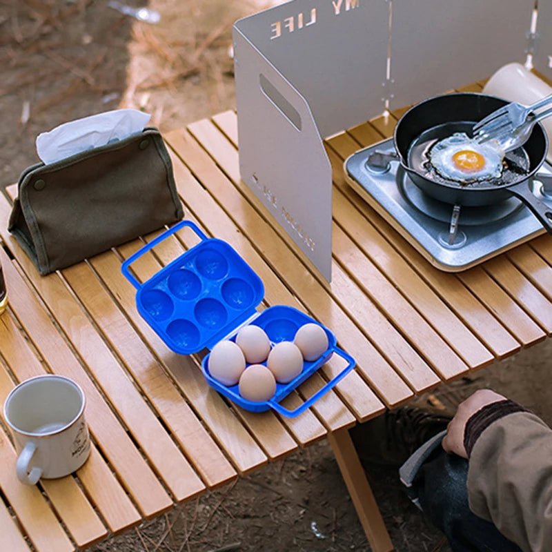 {{Camping Gear {{ Hiking Gear }} }} - {{Tac Treasure Shop }} {{ Blue 6 pack case on wooden table. Single burner grill with cast iron pan with a fried egg cooking. Wind protector set up around burner. Napkin holder and coffee cup . You can see a persons hand in front of table.   }}