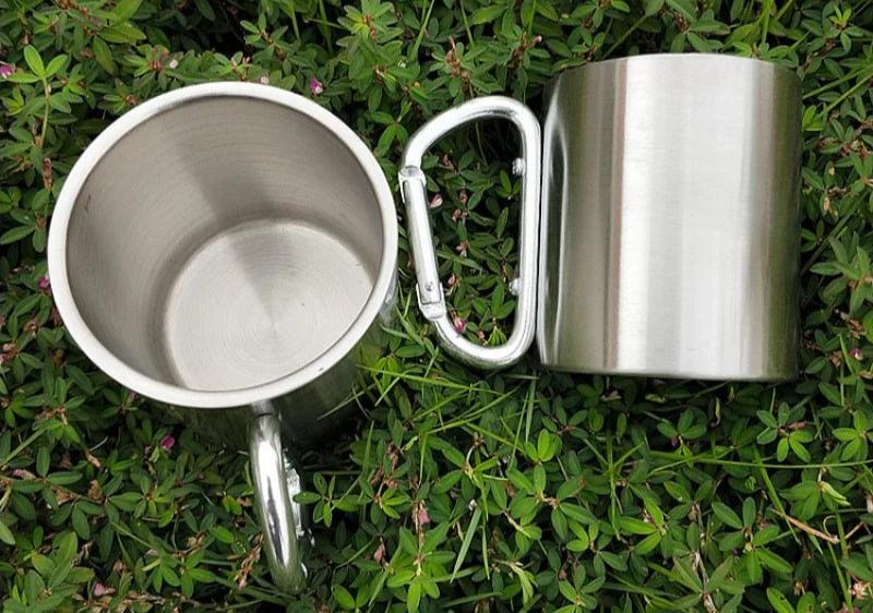 {{Camping Gear {{ Hiking Gear }} }} - {{Tac Treasure Shop }} {{ Two silver carabiner clip handle  stainless steel mugs, shown on top of greenery. One is upright to see inside mug.  The other is on its side to fully display carabiner clip handle. }}